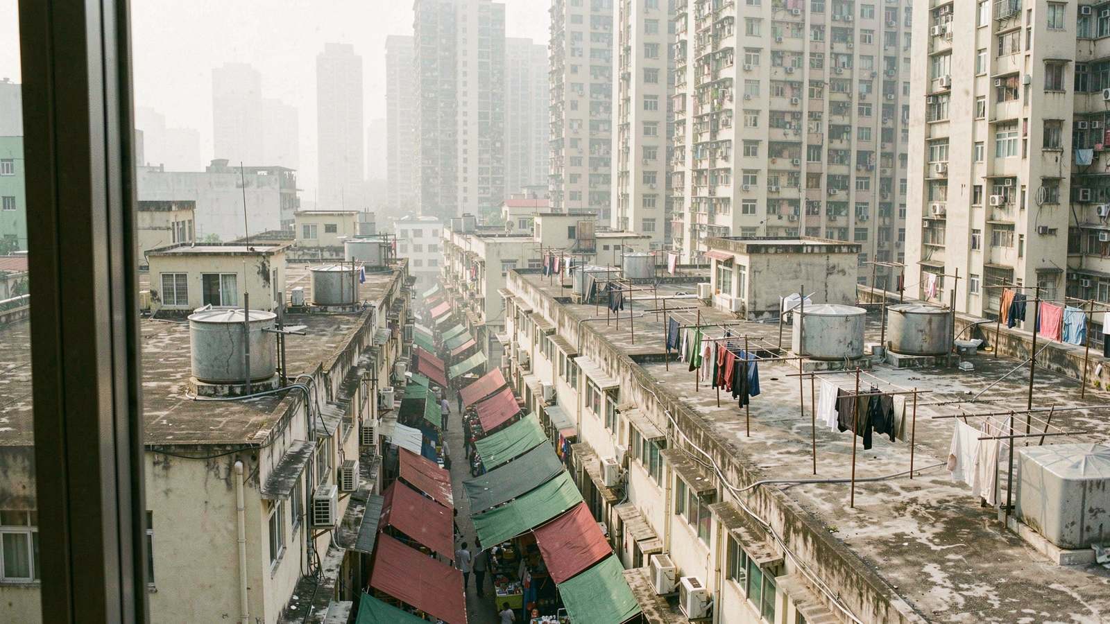 Early morning view from a high hotel window over the crowded rooftops of old Kowloon tenement blocks in Yau Ma Tei, rooftop water tanks and drying laundry, dense residential towers rising behind in soft haze